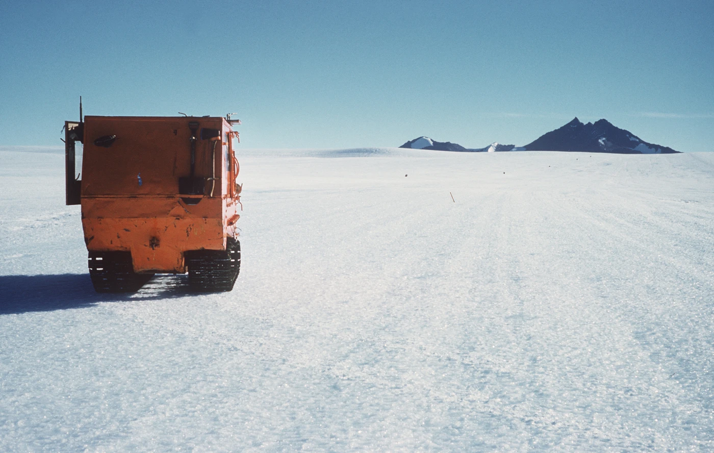 Rear view of a bright orange tracked vehicle in a vast, icy landscape, with rocky mountains on the horizon.