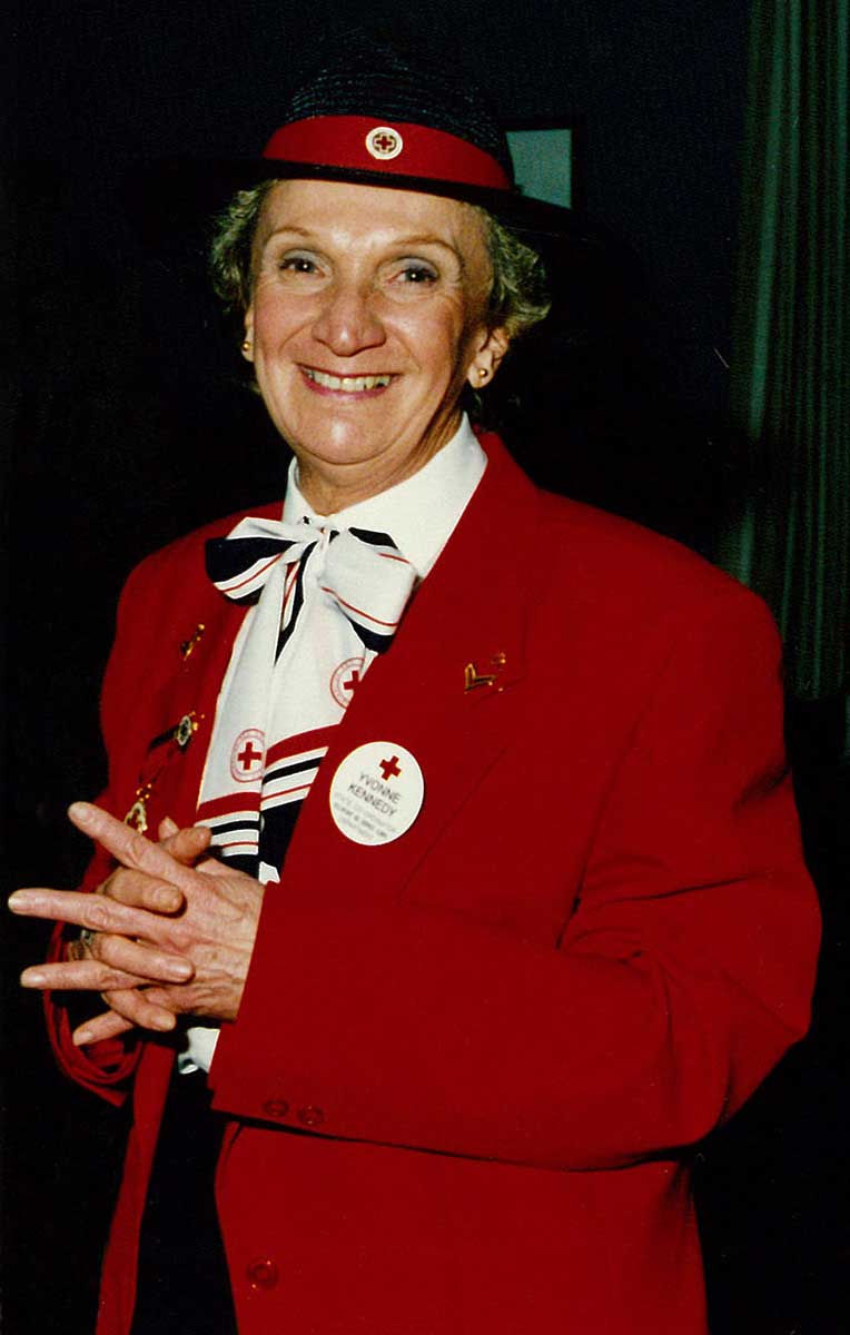 Portrait showing a woman wearing a red jacket and a black hat with red band and pin. The woman is smiling and wears several Red Cross badges and pins. - click to view larger image