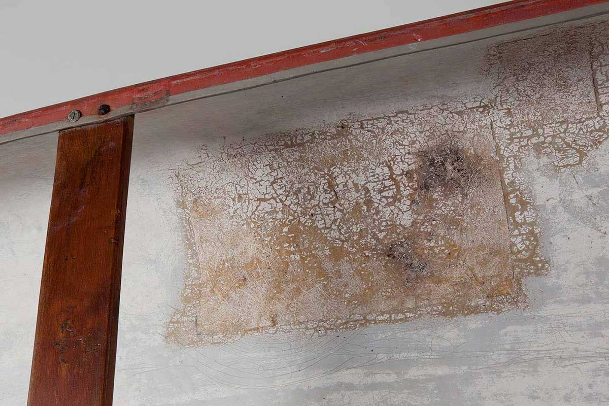Detail of a white surface with worn and degraded paintwork. A wood plank is screwed to the edges of the object. - click to view larger image