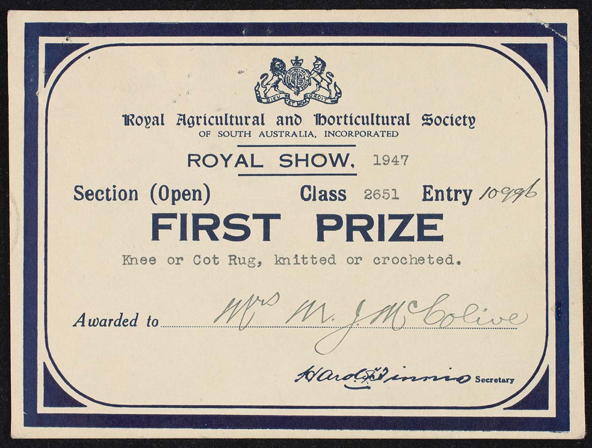 A photo of a first prize card from the 'Royal Agricuttural and Horticultural Society of South Australia', awarded to 'Mrs MJ McColive'  for 'Knee or Cot Rug, knitted or crocheted'. - click to view larger image