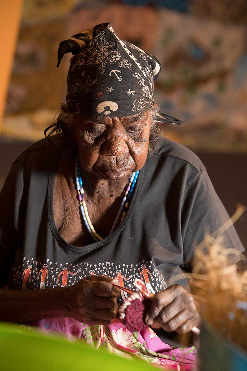 An older woman, adorned with head scarf and beaded necklaces, knits in the 'Songlines' arts hub.