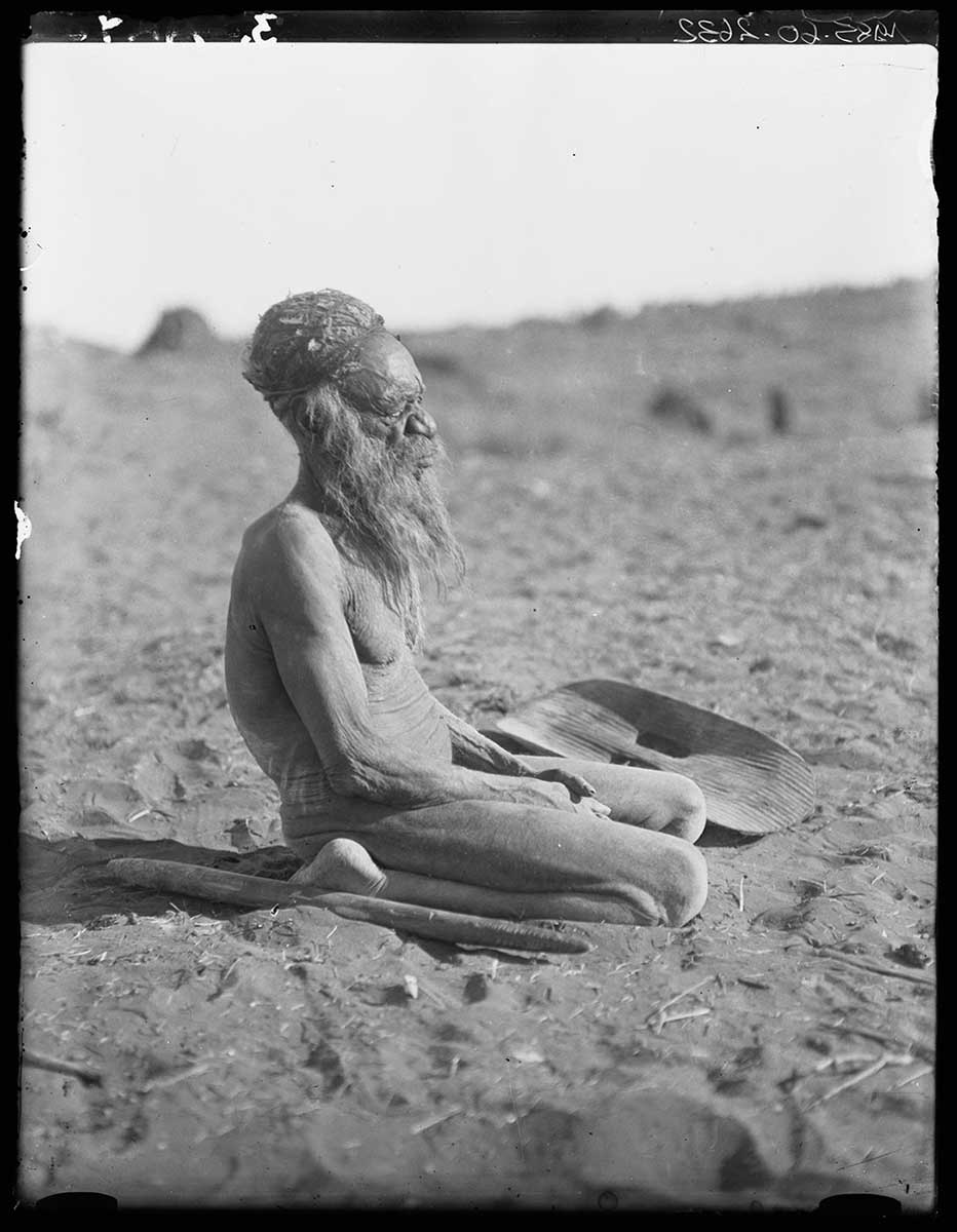 An elderly Aboriginal man, Henbury station, Northern Territory. He sits on his haunches on the bare, fine soiled ground, facing the low sun beyond the right of the image. He wears a close fitting cap made from emu feathers and human hair string. A customary wooden shield as at his left side while a digging stick lies at his right side. The out of focus background has some scattered boulders leading to the horizon which slopes from right to left.  - click to view larger image