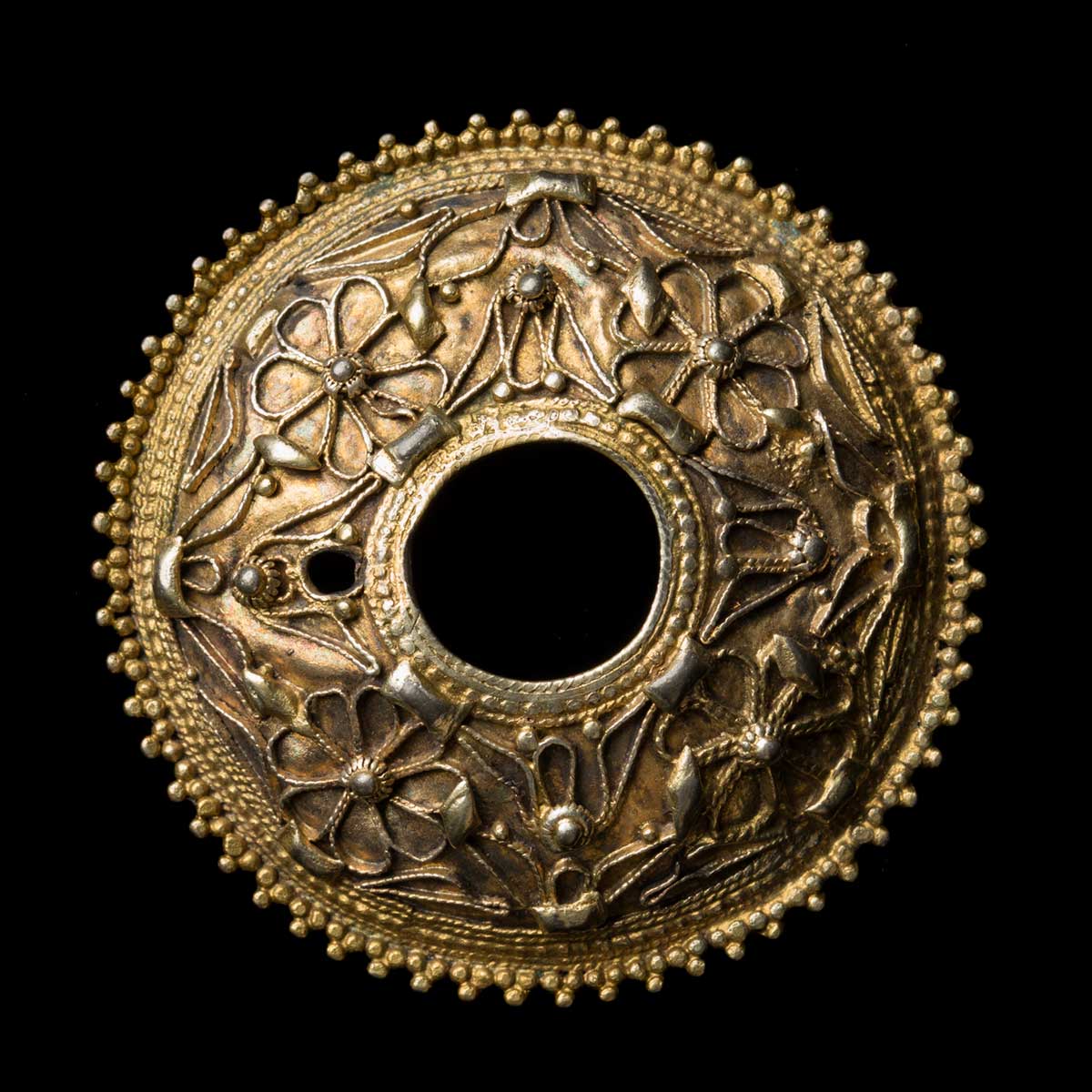 A gold-coloured round button brooch. The exterior edge has intricate beaded details, and the interior edge is decorated with a thin gold-coloured band. The face of the button brooch is decorated with gold-coloured wire details in floral shapes. - click to view larger image