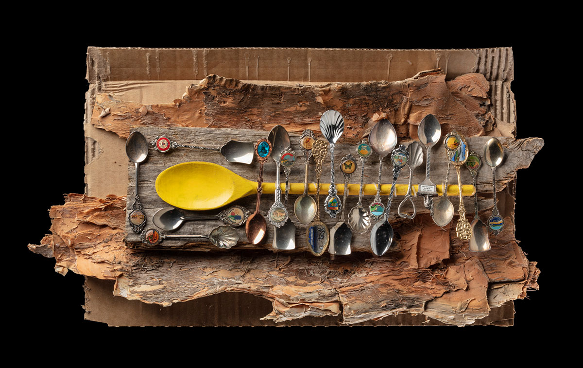 An artwork comprised of a yellow wooden spoon mounted to a piece of grey coloured timber. Adhered to the timber around the spoon and to the handle are twenty souvenir teaspoons. The timber is attached to a piece of paperbark which is adhered to a section of cardboard.