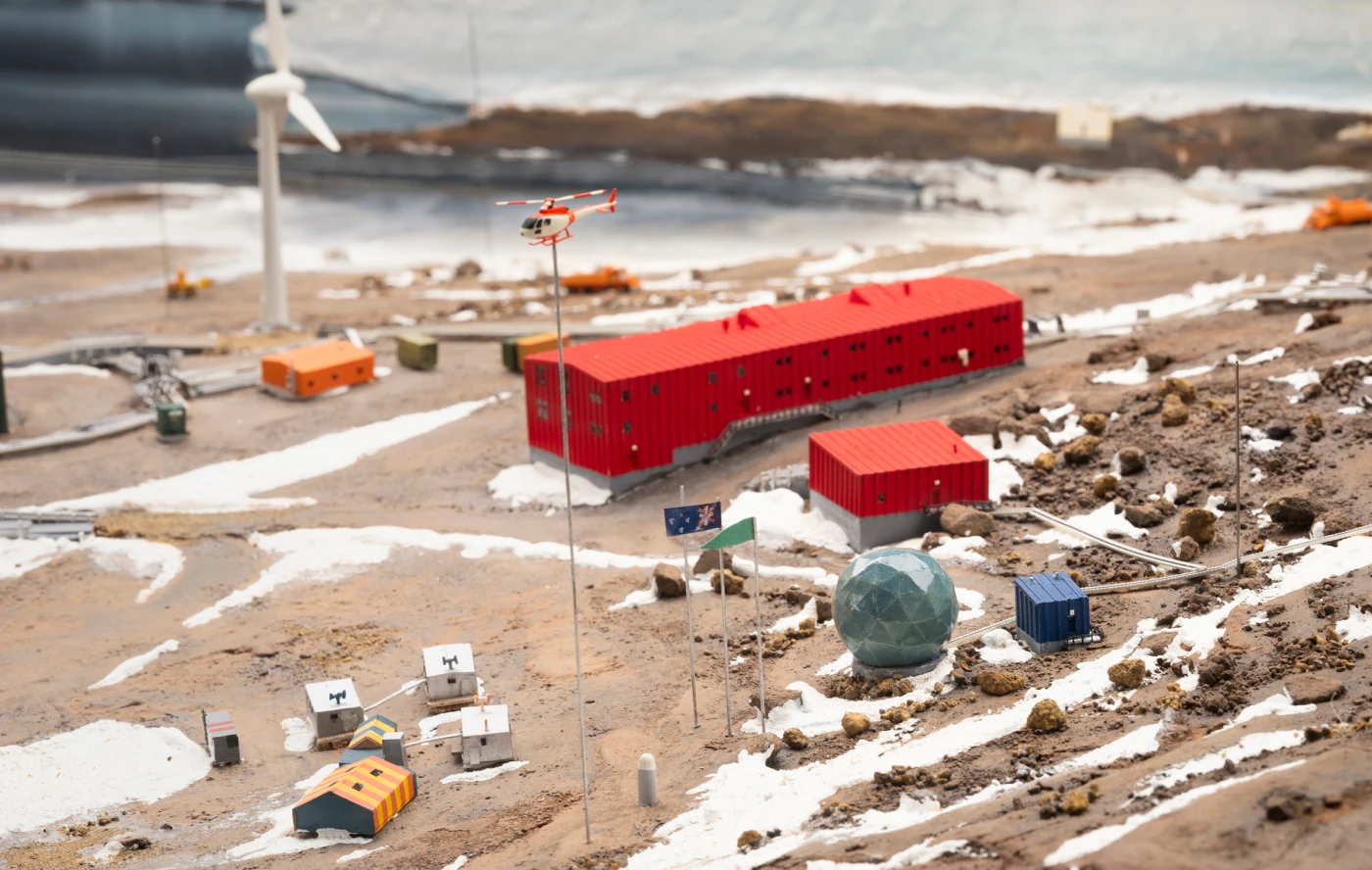 Model of an Antarctic station showing a large red, three storied building at centre, with various outbuildings, a model wind turbine and a small helicopter raised on a stick above. The model terrain is a mix of rocks and snow.