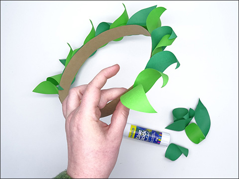 Fingers of one hand grip one part of a headband. The headband is adorned with green paper leaves. There is a glue stick and several pieces of green curled paper to the right of the hand.