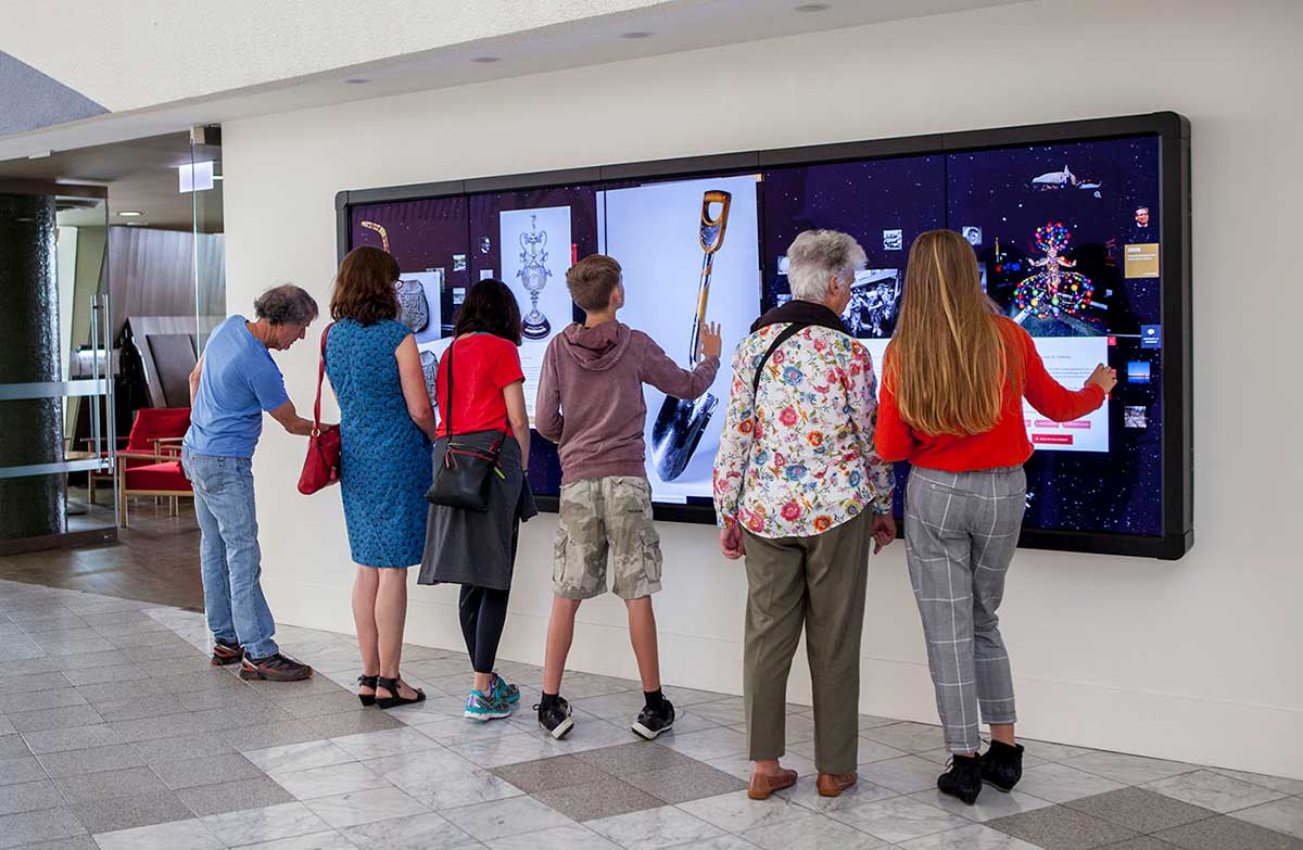 Group of people facing a large, digital screen covered with images and text.