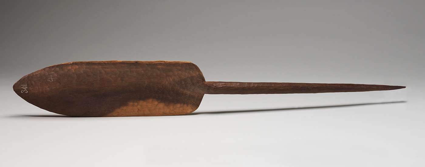 A wooden digging stick with a slightly concave blade narrowing to a cylindrical handle which tapers to a blunt point.