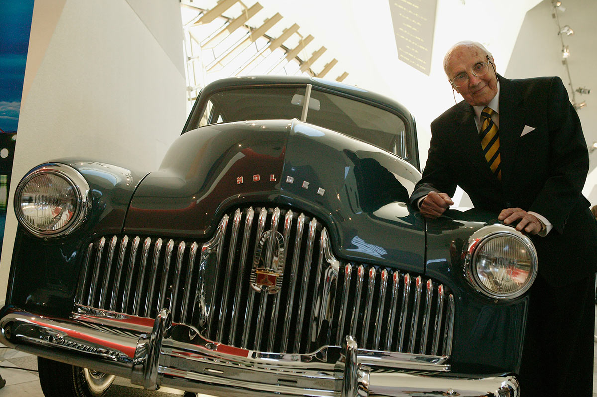 Jack Rawnsley leans on the front righthand side bonnet of a dark blue Holden. The silver grille and headlamps are prominent. The Museum's Hall forms a backdrop. - click to view larger image