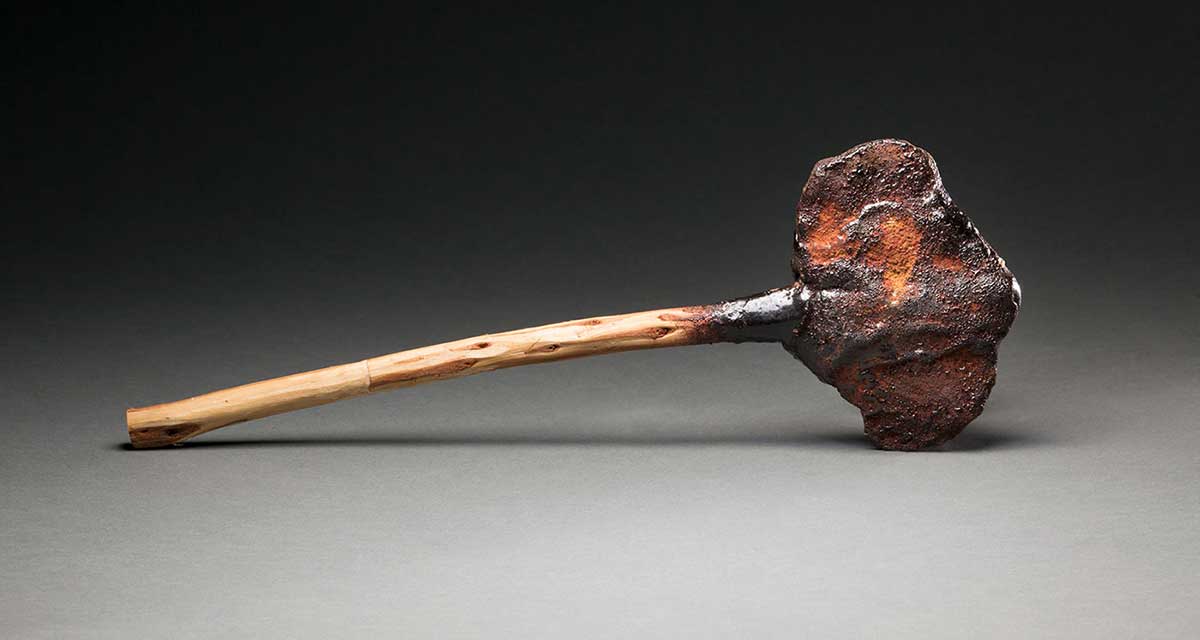 A 'Kodj' axe with a stone head and a wooden handle. The handle is connected to the head with dark coloured resin. The head of the axe has a rust coloured substance on it which may be part of the resin.