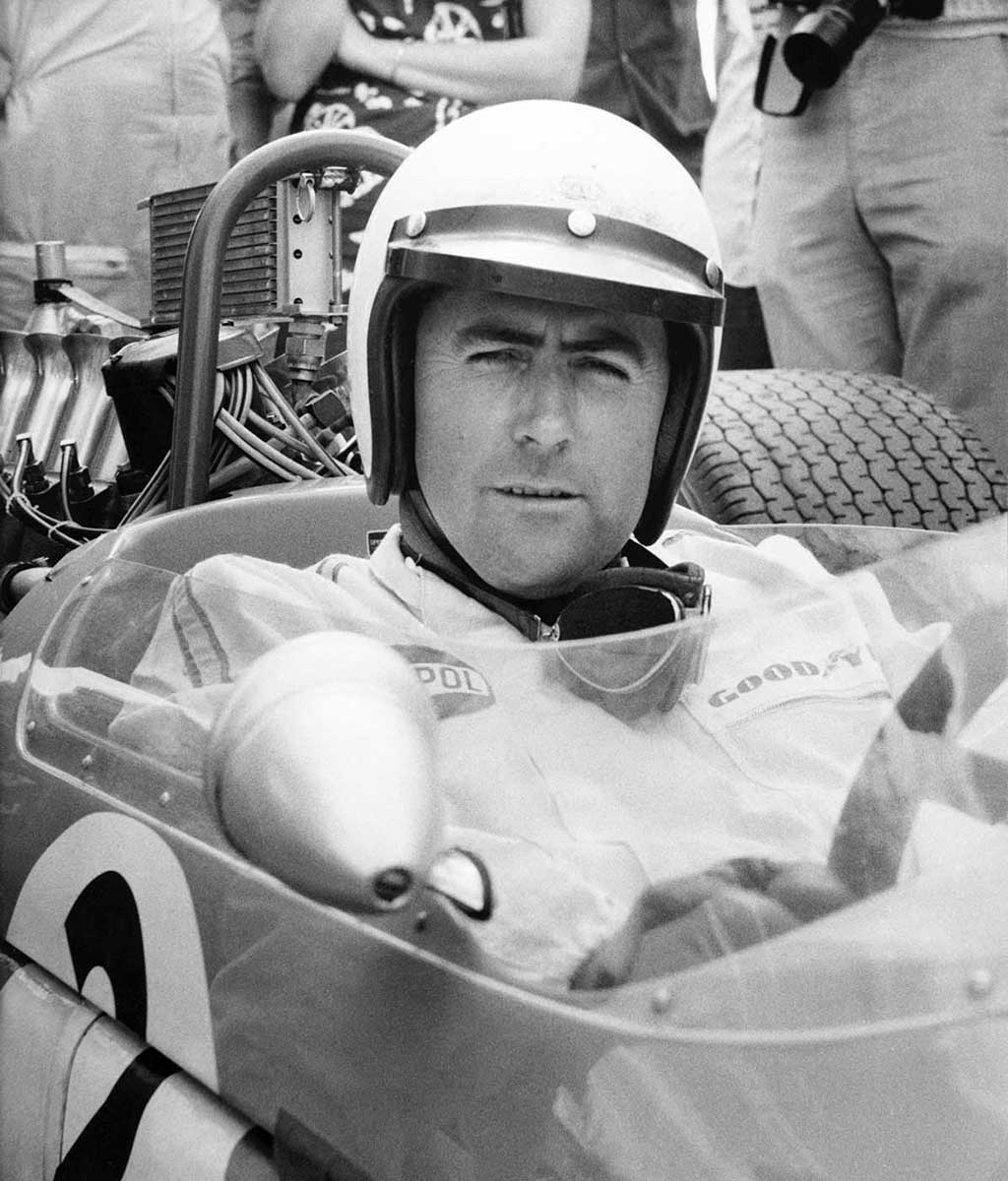 Black and white photograph of a man wearing a helmet in race car. - click to view larger image