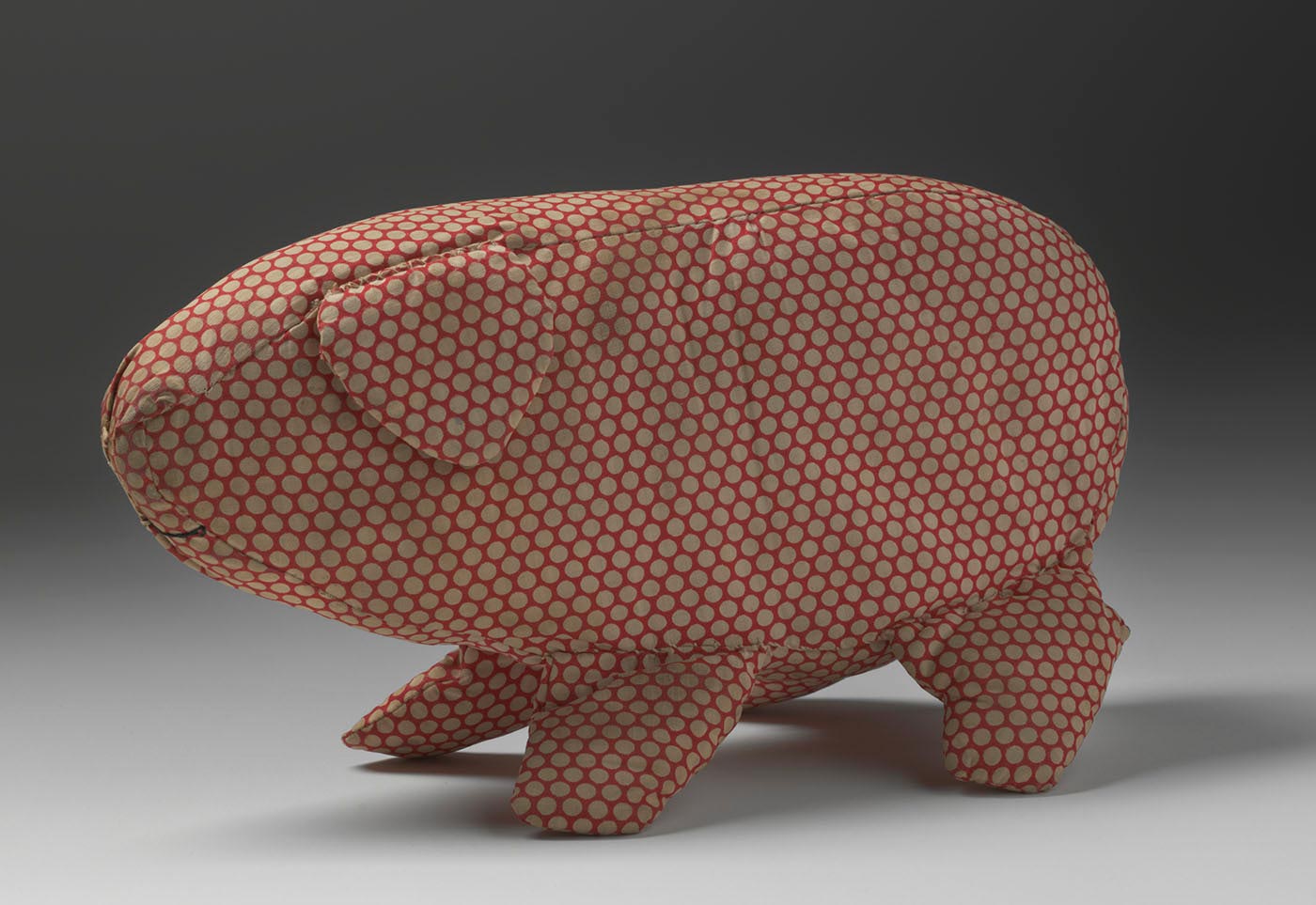 A soft toy in the shape of a pig made from off-white spotted red cotton fabric. The pig has a triangular ear and is missing its tail. A curved mouth is sewn onto the fabric in black thread. - click to view larger image