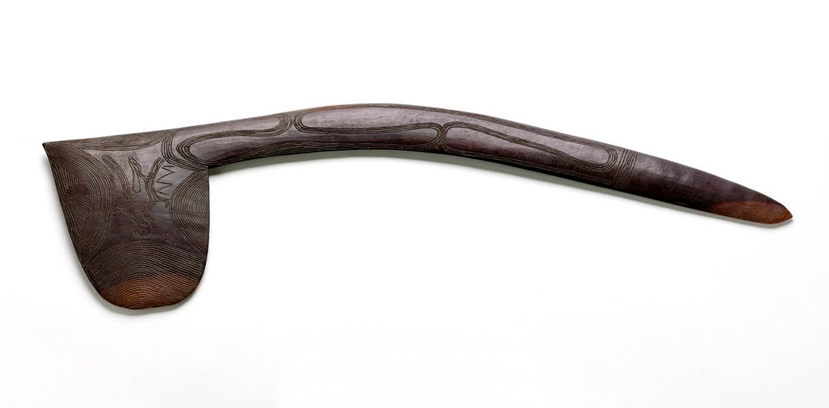 A wooden club with curved handle. A human form has been engraved on the striking section and patterns are visible on the handle.