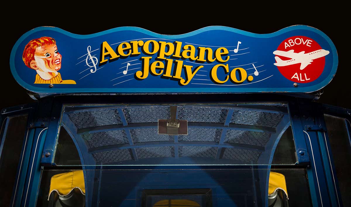 Top section of the front of a truck showing glass windscreen and a blue board painted with text 'Aeroplane Jelly Co', 'Above All' and a whistling boy.