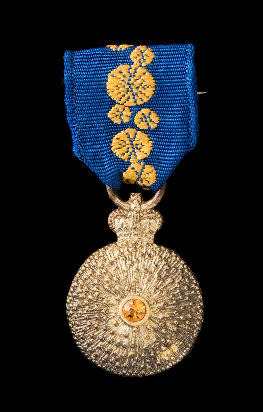 A miniature Order of Australia medal with ribbon. The circular medal has a crown at the top, and is gold in colour with a central amber coloured inset and a raised radiating surface pattern. The medal is hung from a royal blue ribbon with a central band of stylised wattle blossoms, which is attached to a bar clasp pin which is gold in colour. - click to view larger image