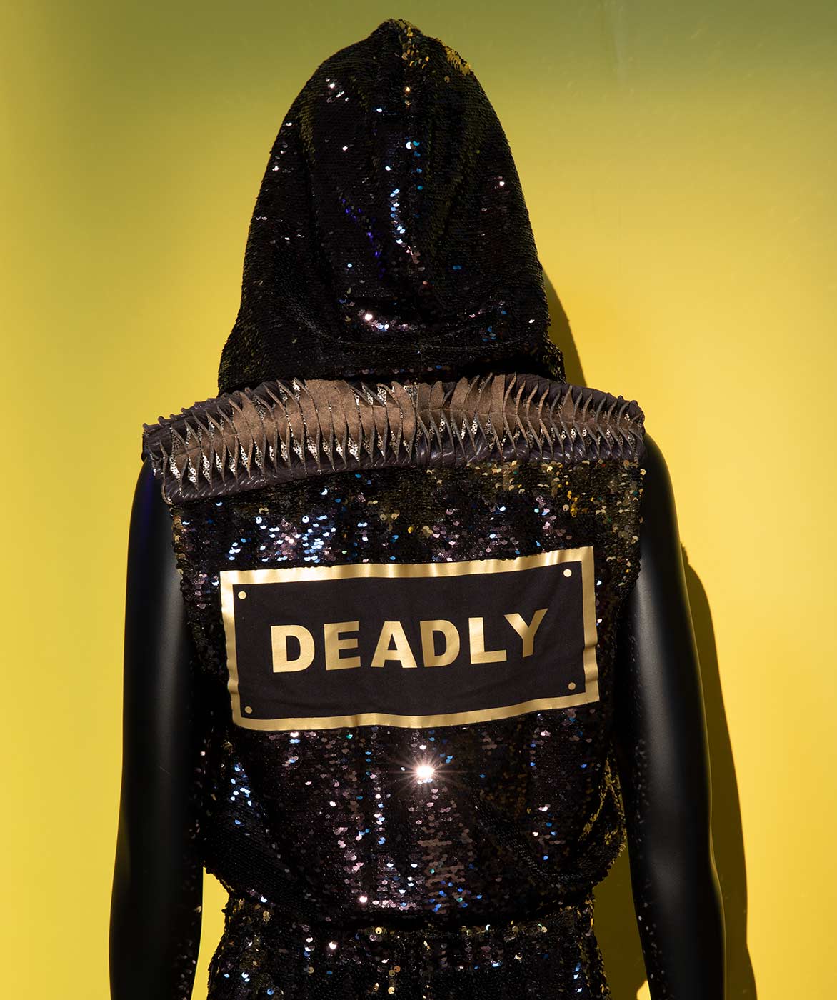 Rear view of a mannequin wearing a black sequinned jumpsuit and hood. On the back, a printed patch says 'DEADLY' in gold letters. - click to view larger image