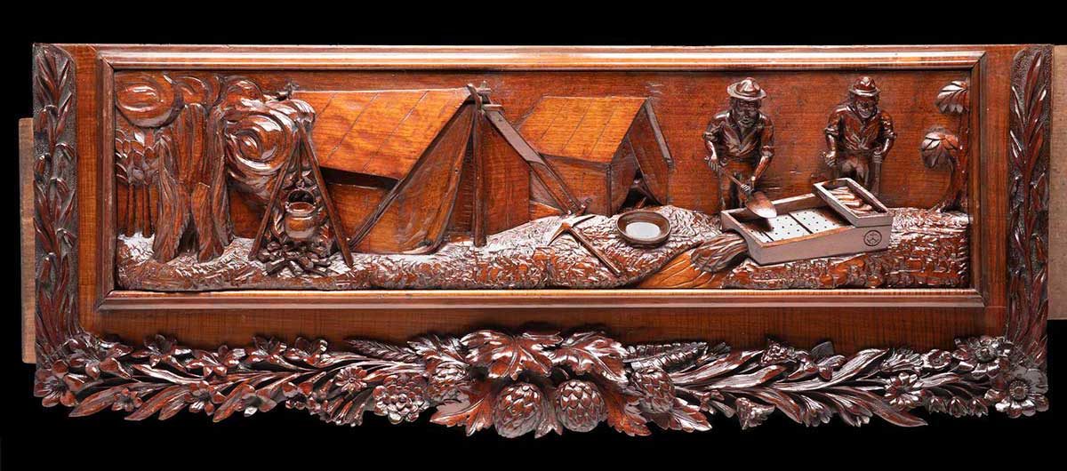 An ornate wooden panel with a relief carving of a scene with two men panning for gold, by a campsite. - click to view larger image