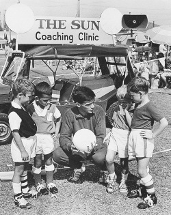 Black and white photo showing a man dressed in a tracksuit, squatting at the same level as four young boys. The man is ressed in a dark tracksuit and boots and holds a white soccer ball. Behind him is a small soft-topped vehicle with a sign on top which reads 'THE SUN / Coaching Clinic'.