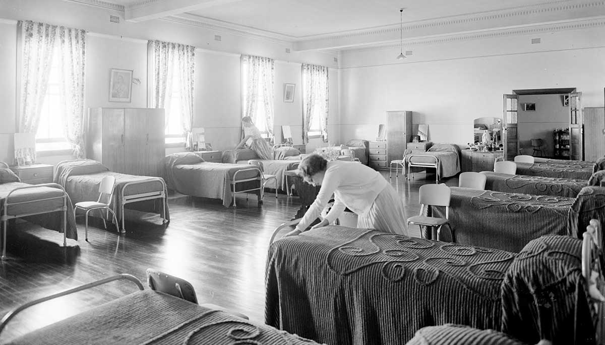 Black and white photo of a girl making a bed in a dormitory.
