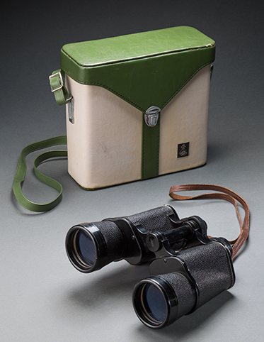 A set of 'YASHICA' brand binoculars, with a black plastic body, and an attached brown leather strap. A 'tasco' brand green and white binoculars case with silver clasp and an attached green shoulder strap.