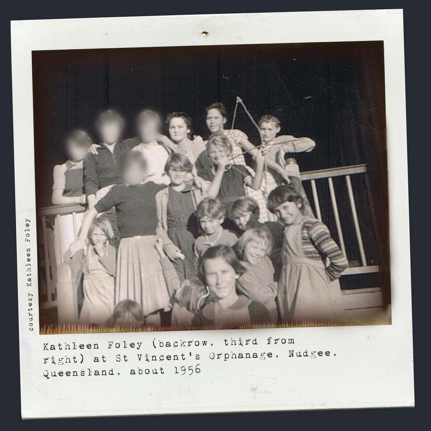 Polaroid photograph showing a group of girls standing at the front of a timber verandah rail. One of the girls holds a small bow and arrow. Some of the older girls' faces have been blurred. Typewritten text at the bottom reads 'Kathleen Foley (back row, third from right) at St Vincent's Orphanage, Nudgee, Queensland, about 1956'. 'Courtesy Kathleen Foley' is typed along the left side. - click to view larger image