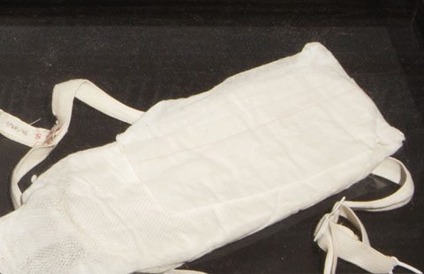 Colour photography of a sanitary pad made of absorbent material with a paper cover. A white cotton and elastic belt is also partially visible. - click to view larger image