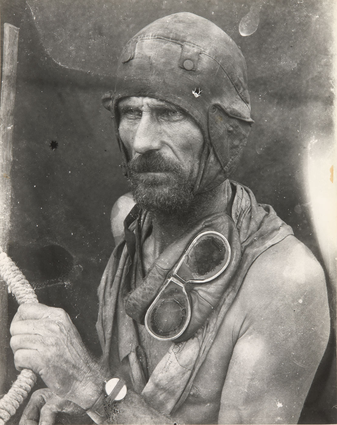 Black and white image showing a bearded and bare-chested man gazing to the left of frame. He wears a leather flying-style cap and has goggles draped around his neck. His right arm clutches a rope. - click to view larger image