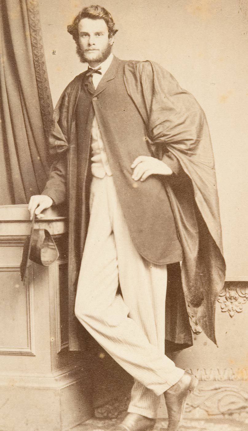 Man posing for photograph in academic gown leaning with right arm resting on a cabinet, with right leg crossed in front of leg and right shoe tip touching the ground and with left hand on waist. - click to view larger image