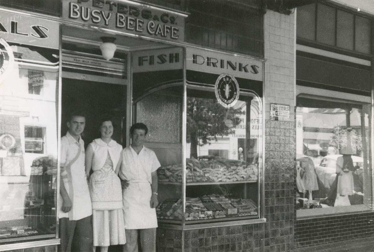Black and white photograph of Mick Georges and an unnamed waitress and cook standing at the entrance of the Busy Bee Cafe.