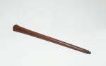 Club made of brown wood. The upper end is larger and rhomboid-shaped – the lower end of the handle has a small knob.