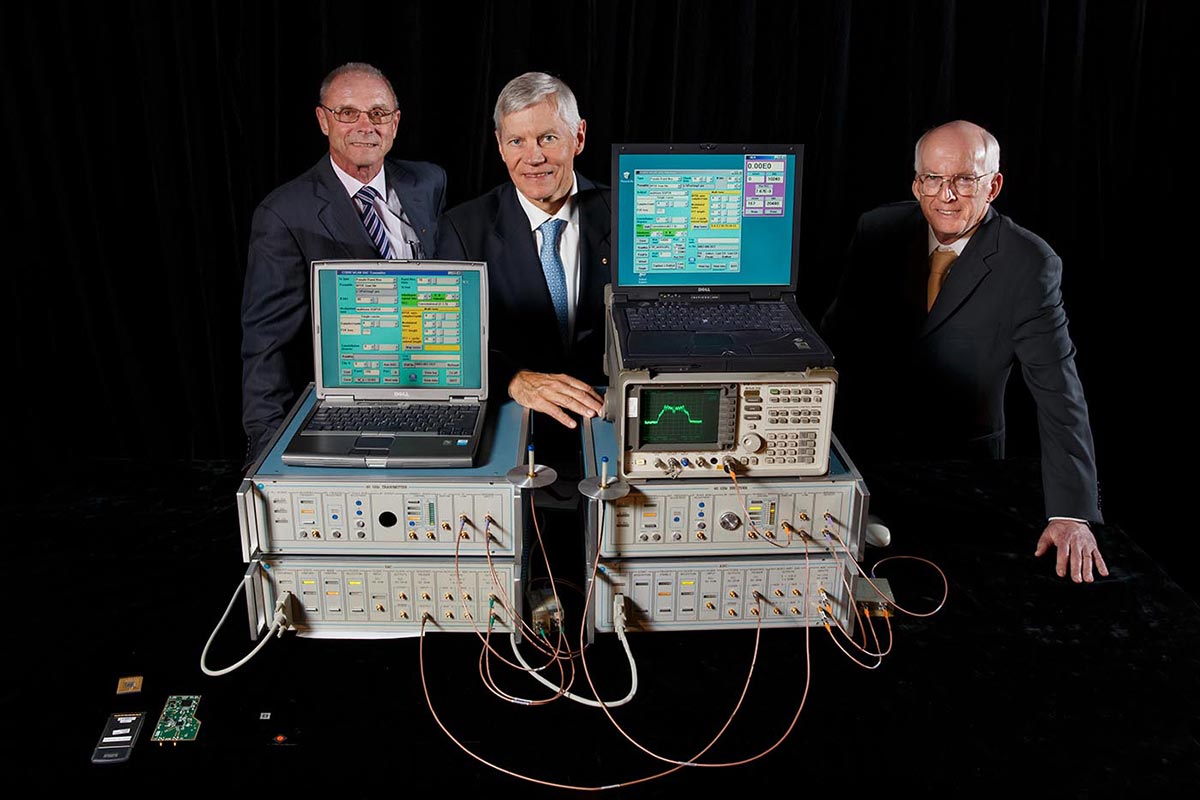 Three men in suits standing beside a table on which sit a collection of cumbersome electronic equipment.