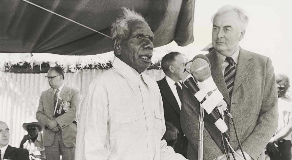 Aboriginal man holding document speaks in front of microphones on stand, with Prime Minister Whitlam to his left.