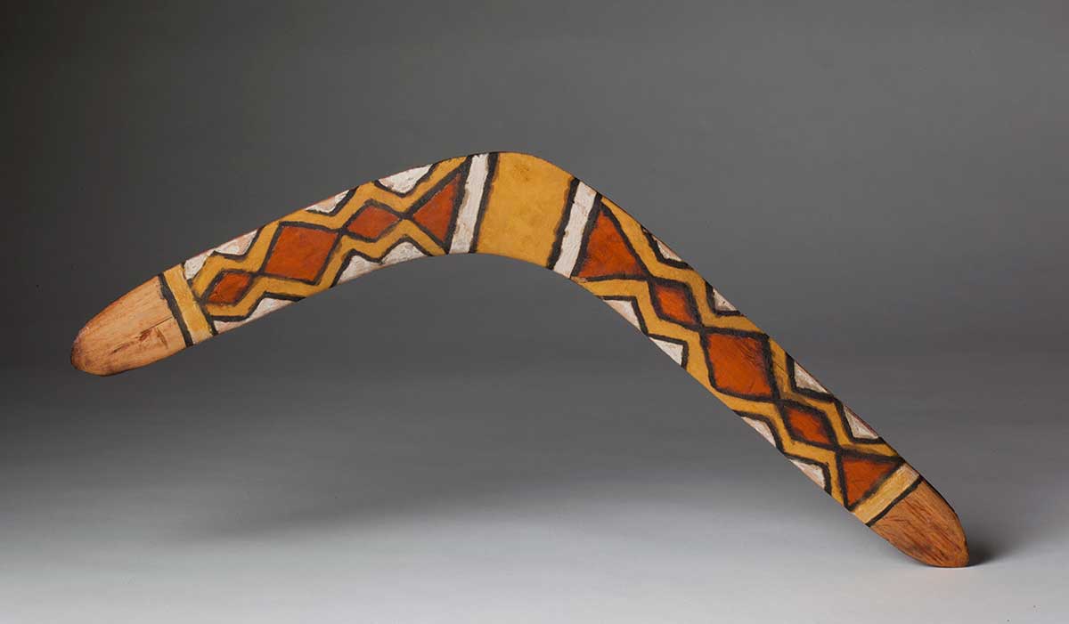 Studio photograph of a boomerang with a painted design consisting of diamond shapes. 