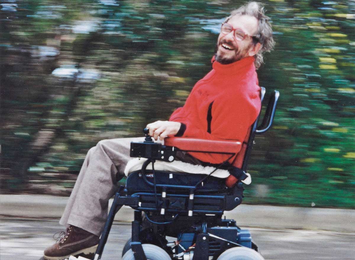 Photograph showing the side view of a man in a motorised wheelchair. The man is smiling, looking at the camera. The landscape behind him is blurred.