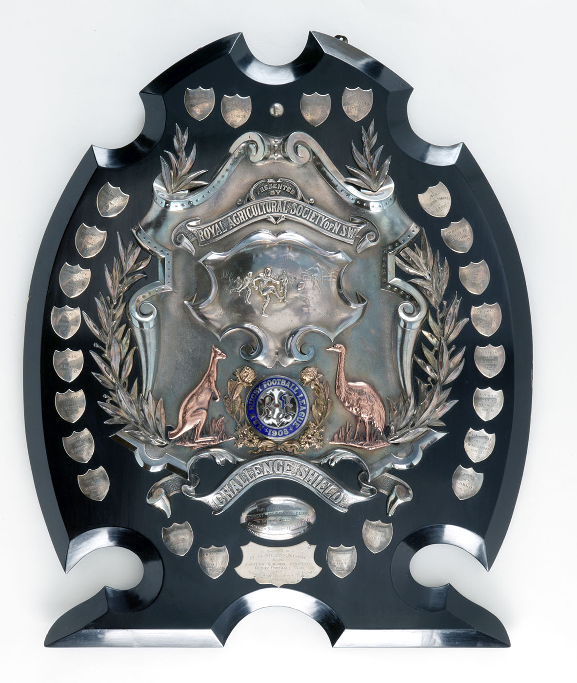 Presentation shield on a timber base. Near the top of the shield, in silver, 'PRESENTED / BY / ROYAL AGRICULTURAL SOCIETY OF N.S.W' is printed on a scroll above a silver relief scene from a rugby match. A gilded kangaroo and emu flank a gilded waratah wreath. - click to view larger image