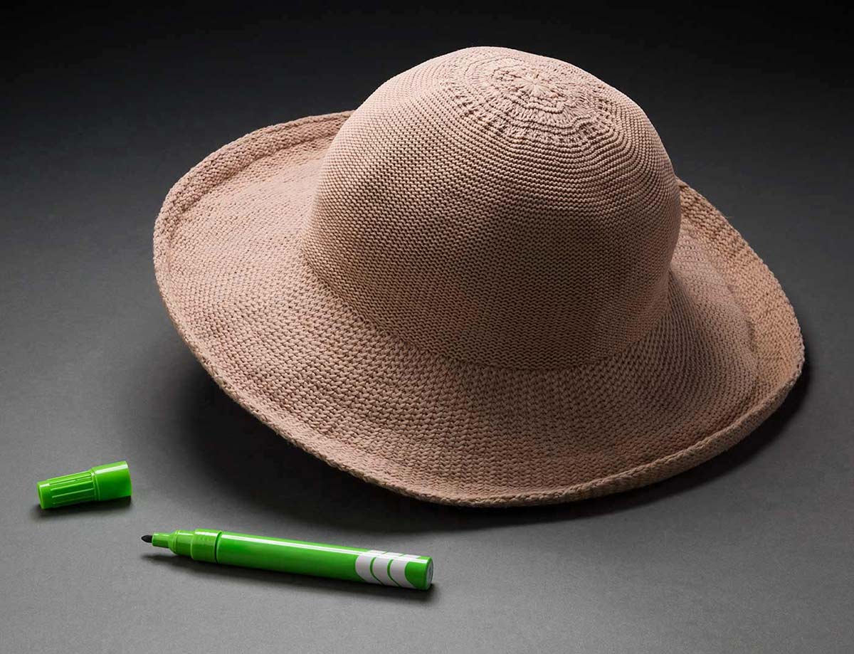 A beige sunhat with a strap and a tag for 'The / Cancer / Council / Australia' attached on the inside of it. A green felt-tip marker pen with the text 'Artline PERMANENT' on one side of it. - click to view larger image