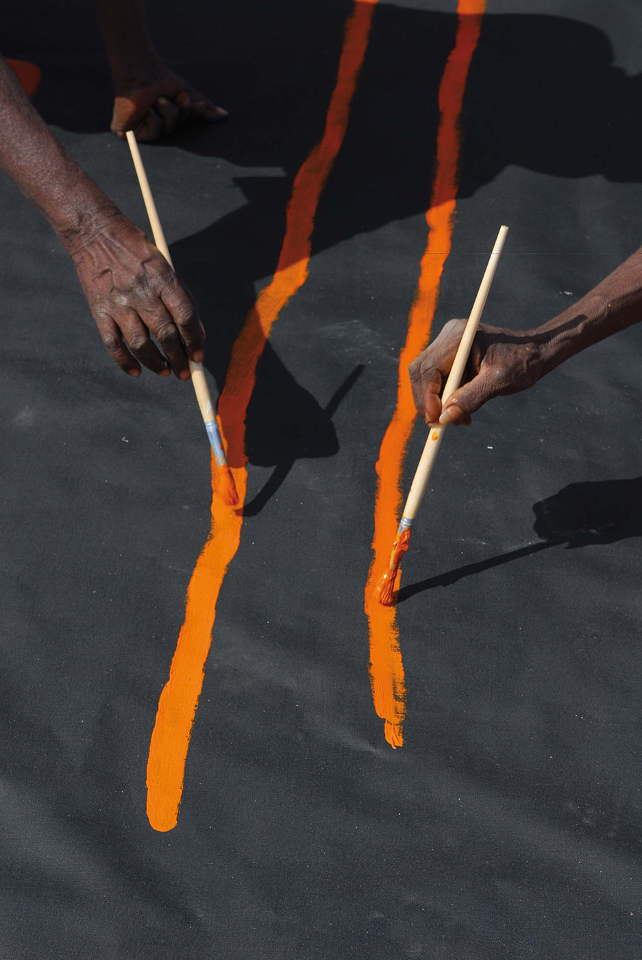 Colour photo of two hands painting orange pigment with onto a canvas. - click to view larger image