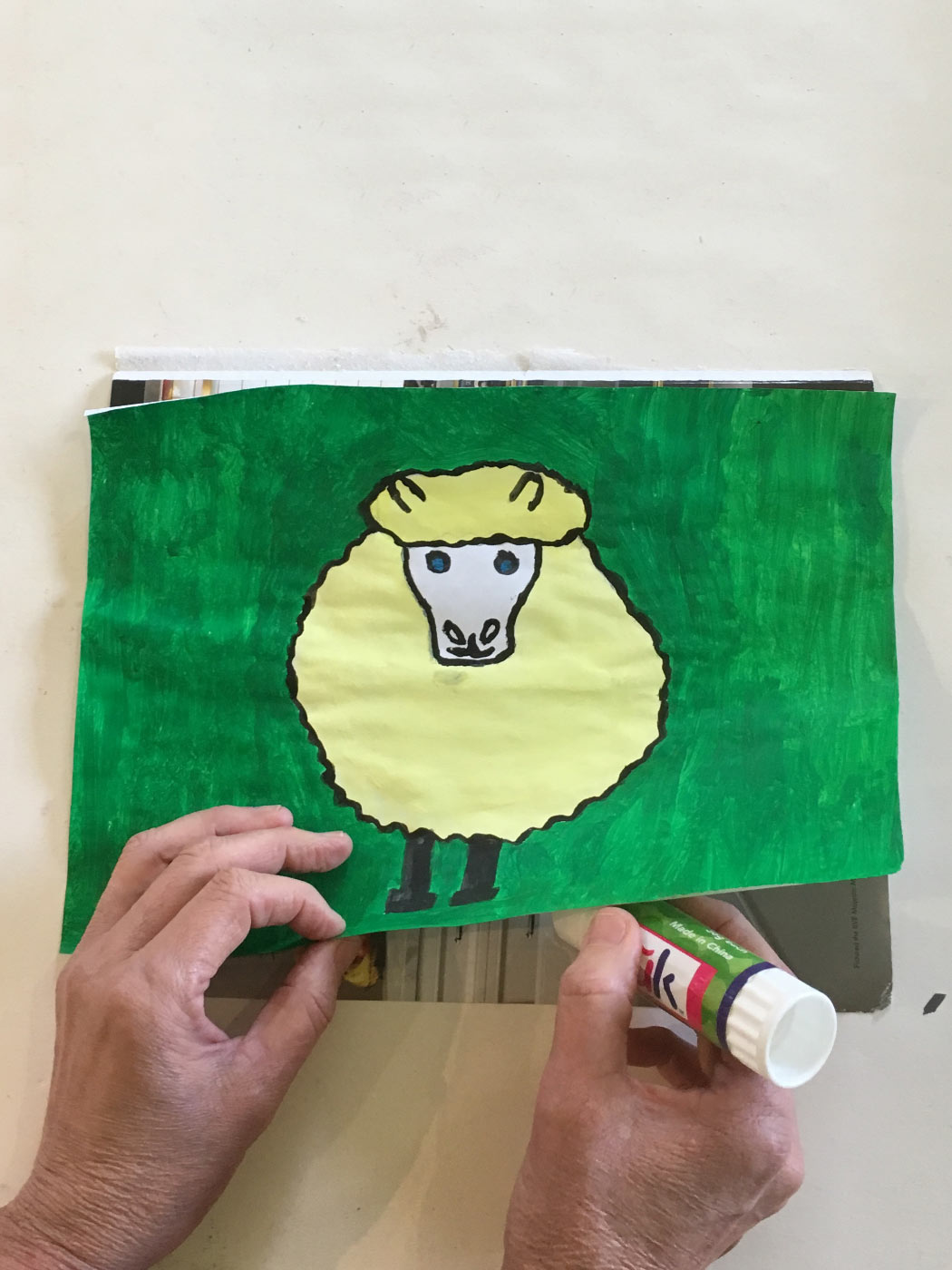 A pair of hands gluing down an illustration of a sheep on paper created with textas and paint.