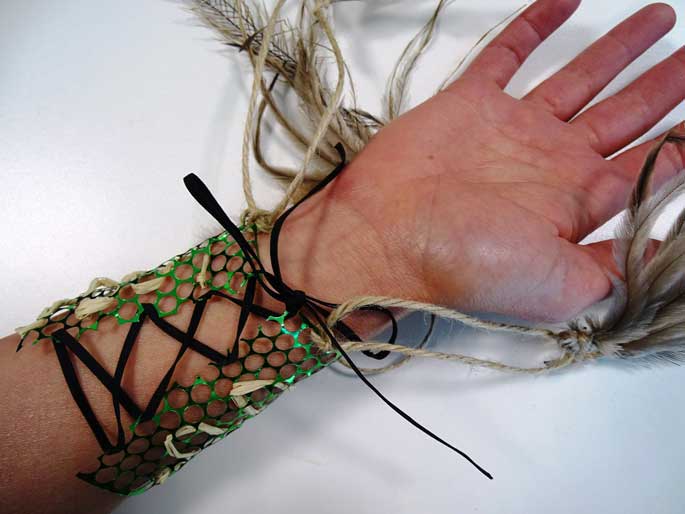 An arm cuff made from a piece of green mesh with elastic.  Feathers are attached to the arm cuff with string.
