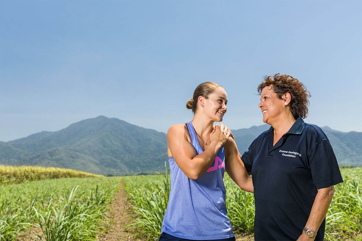 Ashleigh Barty and Evonne Goolagong Cawley against a background of farmland. They are in a half embrace and are smiling at each other affectionately.