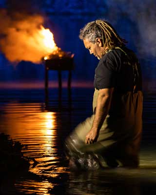 A man in waders and fishing vest kneeling in shallow water. In the distance is a pyre burning against a dark blue background.