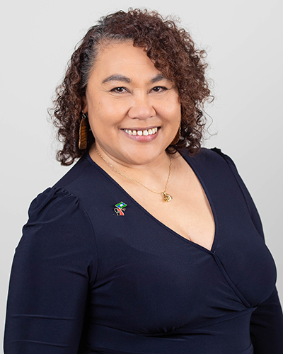 A photograph of Karne Mundine. Karen has shoulder length curly hair and is wearing a dark blue dress with a pin with the Aboriginal Flag and the Torres Strait Islander flag. She is smiling at the camera. 