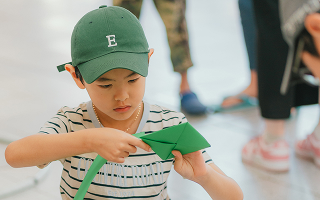 A photograph of a young child playing with a handmade green Korean toy, from a Museum school holiday program. 