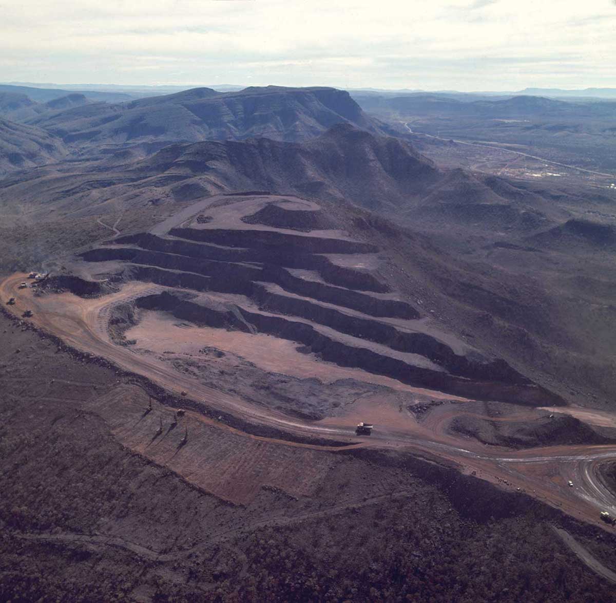 Aerial photo of an open cut mine carved out of the side of a large hill. Massive mining trucks are visible on the perimeter road. Unsullied countryside and mountains in the foreground and background. - click to view larger image