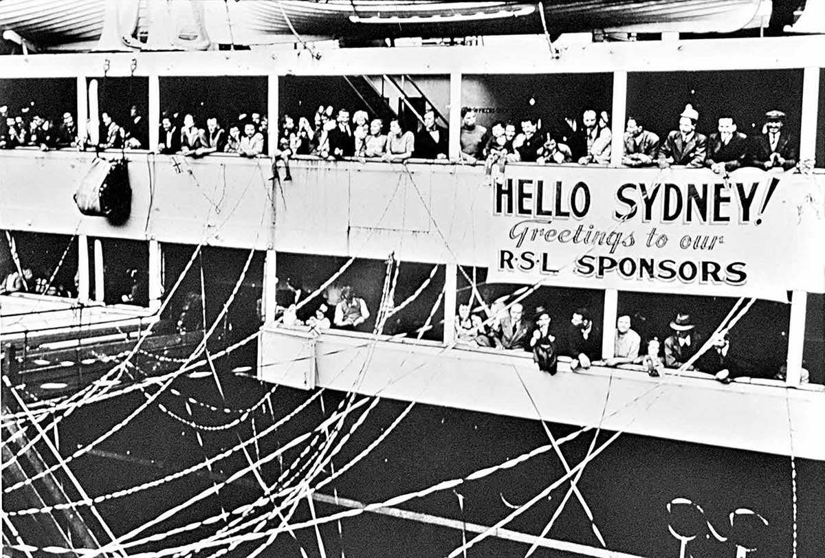 Ship docking with streamers strewn between ship and shore. A banner on the ship reads ‘Hello Sydney!’.