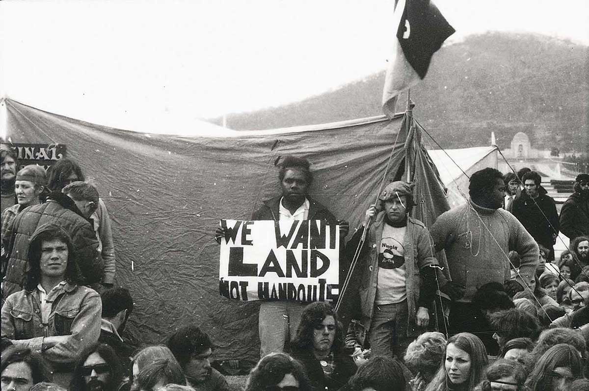 Group of young people, some of whom are Aboriginal, sitting or standing in front of a tent with a flag above it. An Aboriginal man at the centre of the photo is standing up looking at the camera holding a placard that says ‘We want land not handouts’.