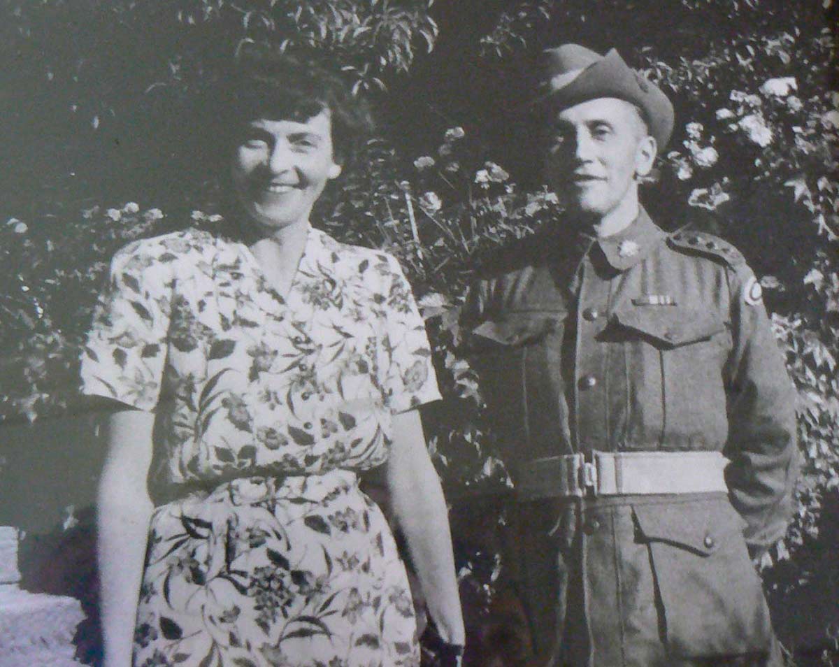 A black and white photograph of a man and a woman. The woman stands at the left of the photo, wearing a dress with a floral print. She has dark hair, and smiles. The man stands at the right of the photo, wearing a Second World War-era army uniform and slouch hat. He has his hands behind his back. Behind them both are large flowering shrubs and bushes.