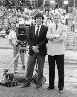 Black and white photograph showing two men standing beside a television camera on a grass field. Both men are wearing suit coats and ties and are crossing their arms. In the background a man wearing a headset uses the camera and there is a crowd of people in tiered seating.