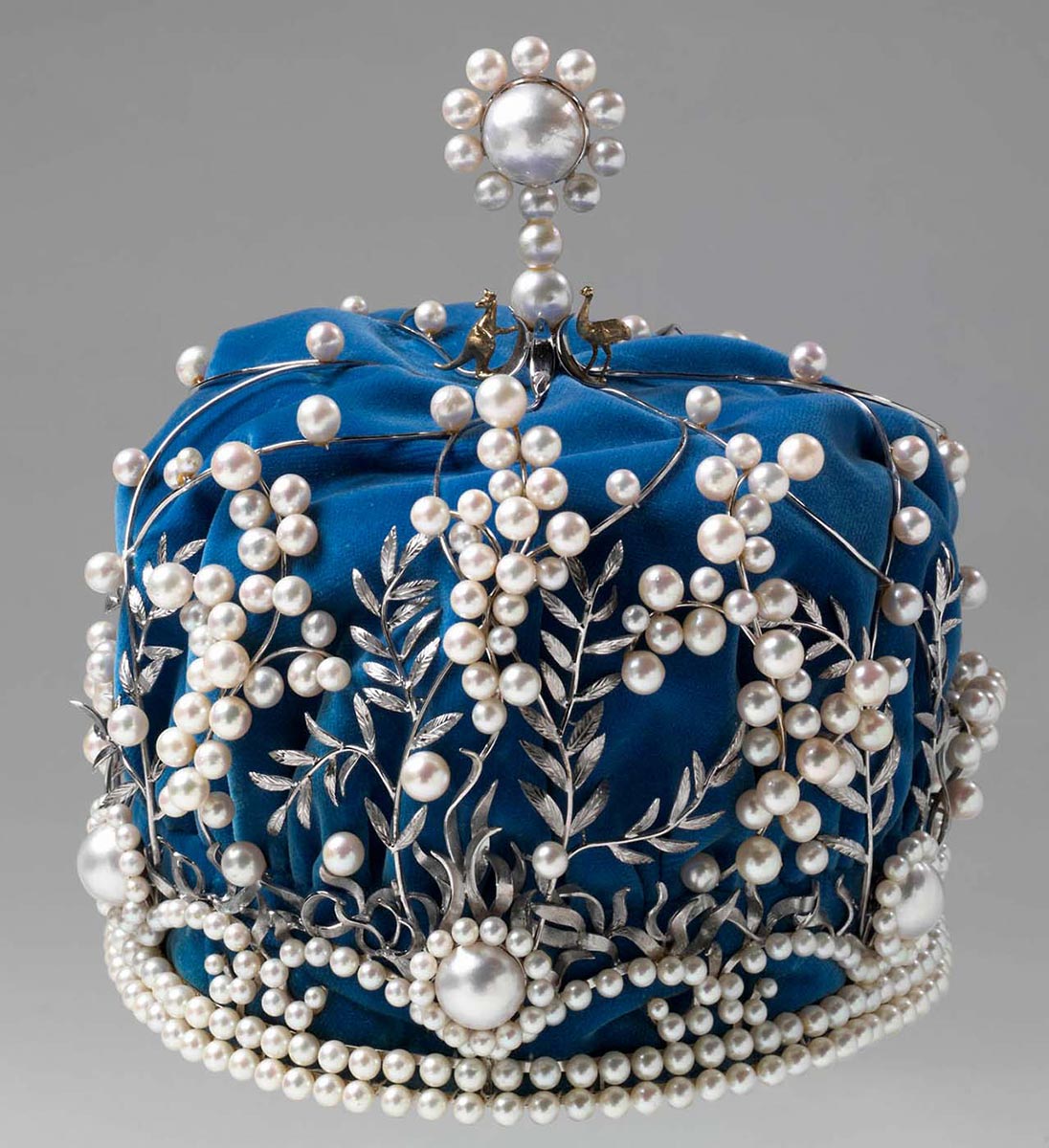 The Miss Australia crown, made with a silver-coloured metal in a wattle-leaf design, inset with cultured pearls. On the top of the crown are small gold-coloured figures of a kangaroo and an emu, standing either side of a upright centrepiece which features a large natural pearl. The crown is lined with blue velvet, which is, in turn lined with white netting.