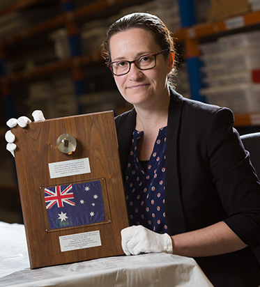 A Woman wearing white cotton preservation gloves, displaying wooden plaque mounted with a preserved object, a small Australian flag and labels printed or engraved with text.